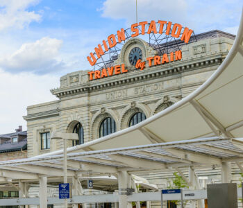 Historic Denver Union Station entrance with iconic Travel by Train sign beneath bright Colorado skies.