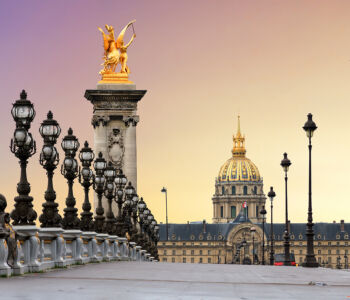 Pont Alexandre III leading toward Les Invalides at sunset, with ornate lamps and a gilded statue in view.