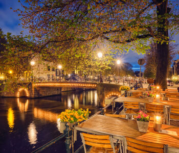 Candlelit canal-side terrace overlooking an illuminated bridge in Amsterdam at dusk.