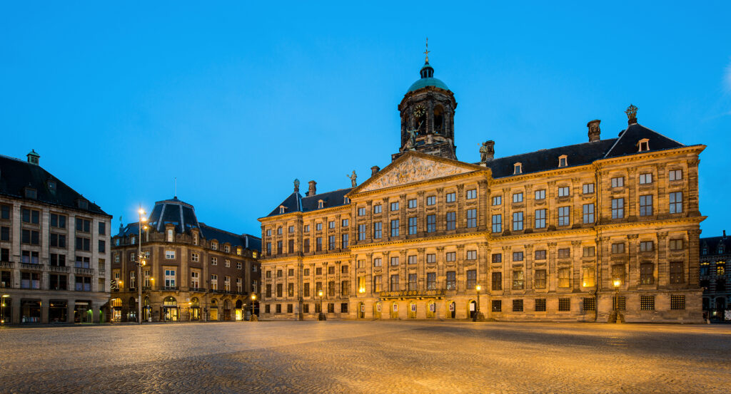 Evening view of the Royal Palace in Amsterdam's Dam Square under a blue sky.