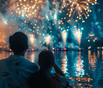 Two people watch colorful fireworks illuminating the night sky over a lagoon at Disney.