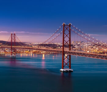 The 25 de Abril Bridge glowing at dusk as warm city lights reflect across the Tagus River in Lisbon.