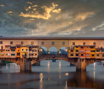 The Ponte Vecchio spanning the Arno River at sunset with warm lights reflecting on the water.