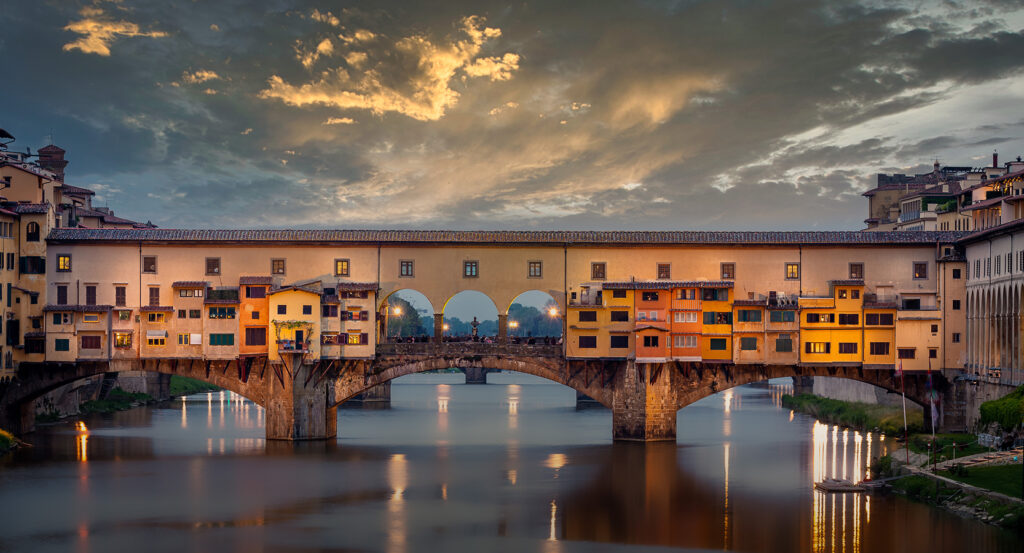 Illuminated Ponte Vecchio with shops and arches reflecting in the Arno River