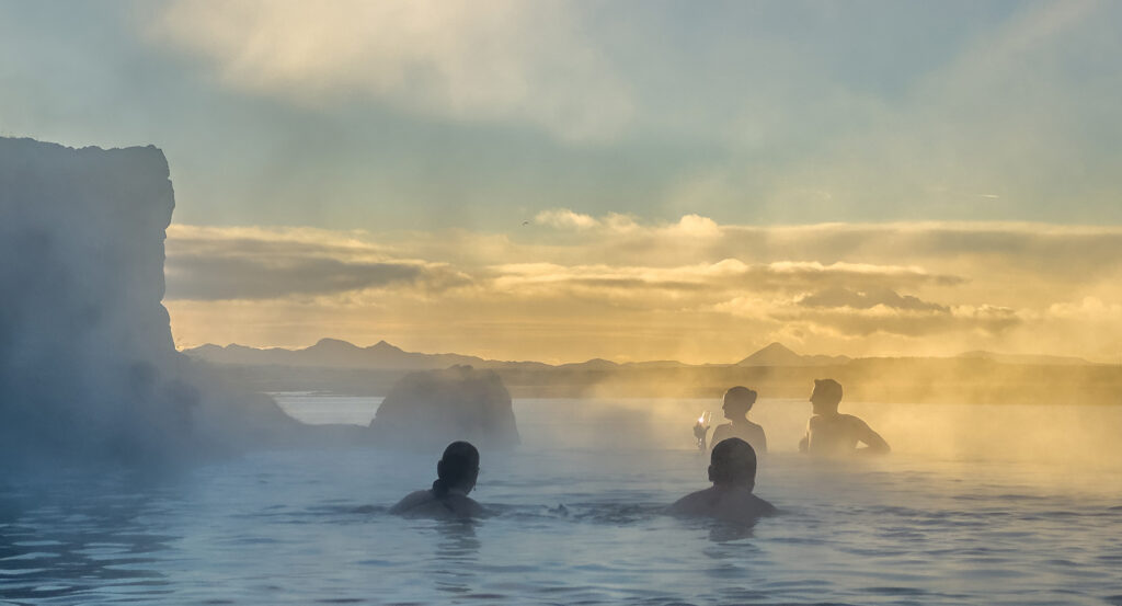 Silhouettes enjoying cocktails in Sky Lagoon's warm waters against golden Icelandic sky and ocean horizon