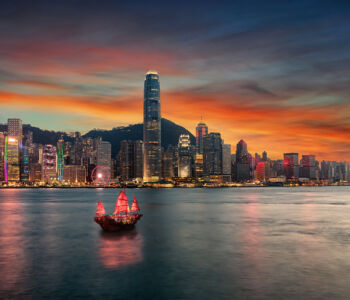 Hong Kong skyline at sunset with a traditional red-sailed junk in Victoria Harbour.