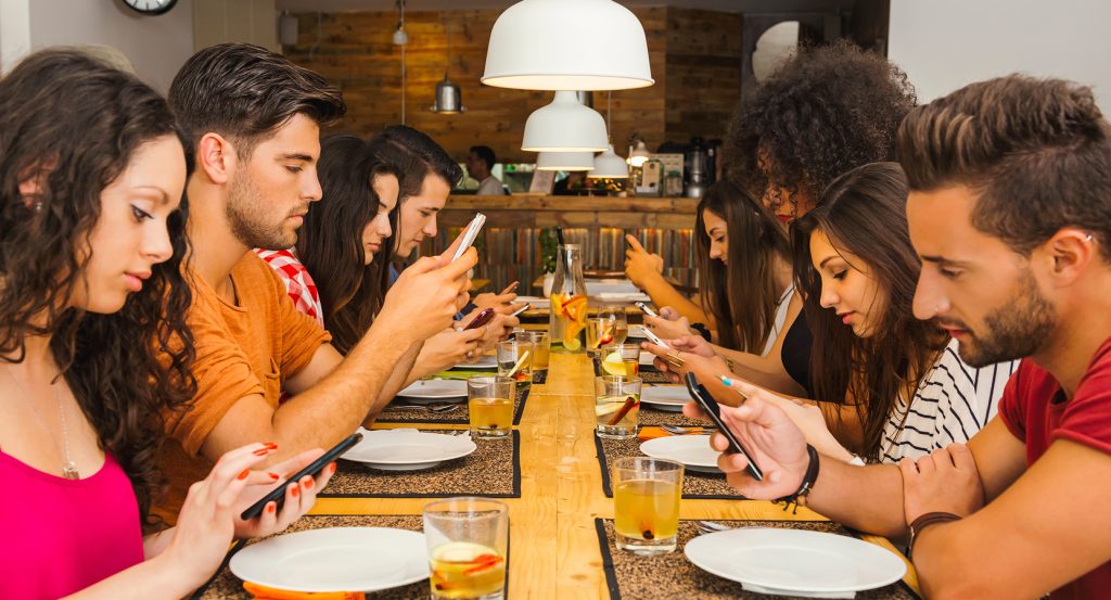 Friends dining together while focused on their phones, highlighting social media’s grip on real-world moments.