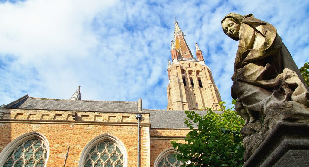 A weathered statue gazes downward beneath Brugge’s medieval spire and cathedral windows.