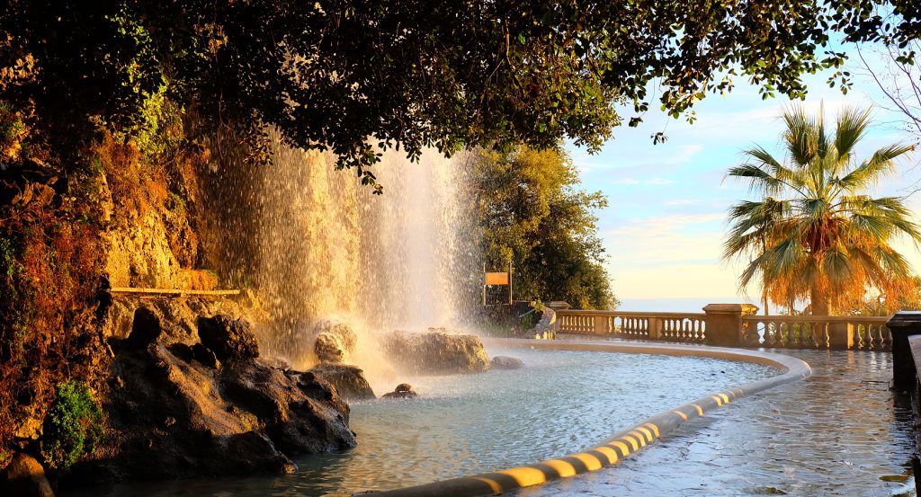Scenic view of a cascading waterfall and palm tree at Castle Hill in Nice during sunset.
