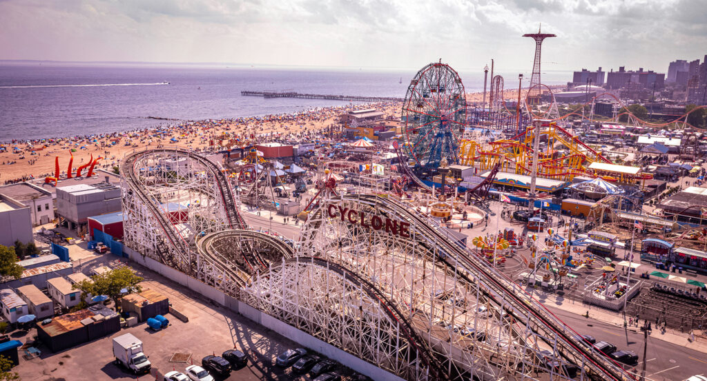 Coney Island boardwalk and beach filled with crowds, with roller coasters and Ferris wheel visible near the ocean.