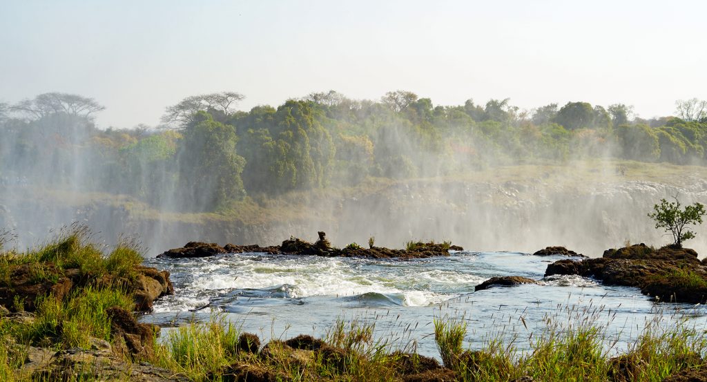 A tranquil rock pool overlooks the thunderous drop of Victoria Falls, framed by wild greenery.