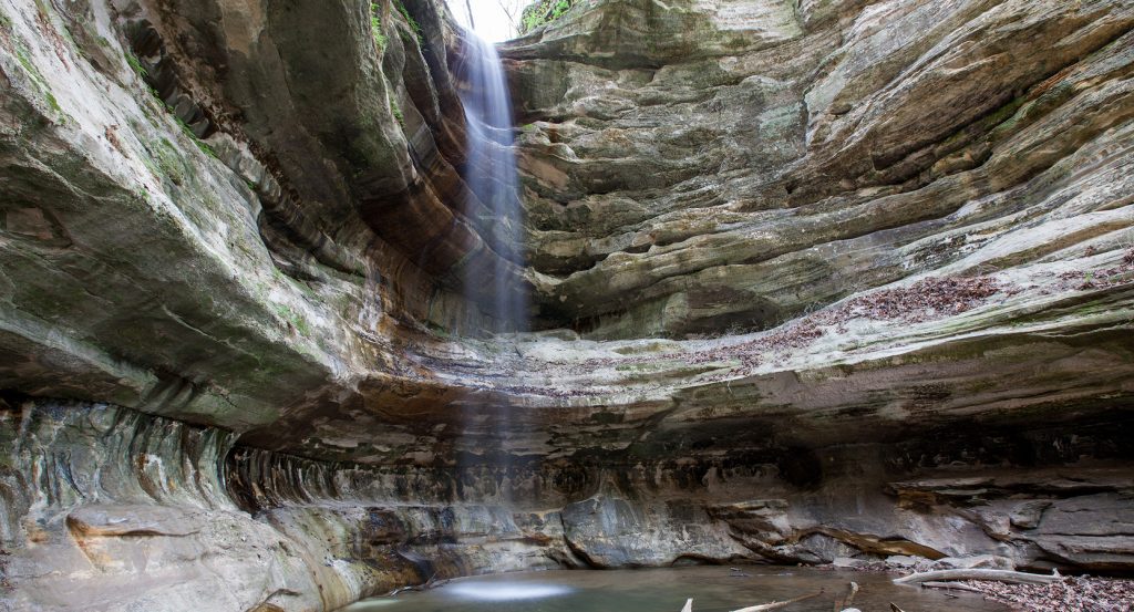 Peaceful cave waterfall flowing into a shallow pool surrounded by driftwood and stone textures.