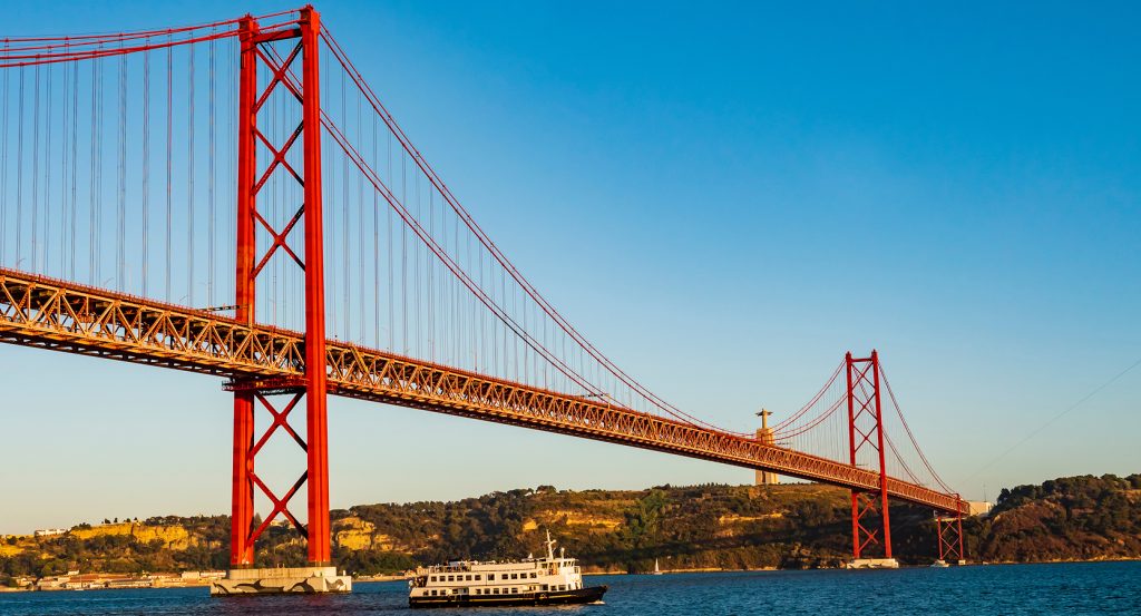 Passenger ferry gliding beneath Lisbon’s 25 de Abril Bridge at golden hour