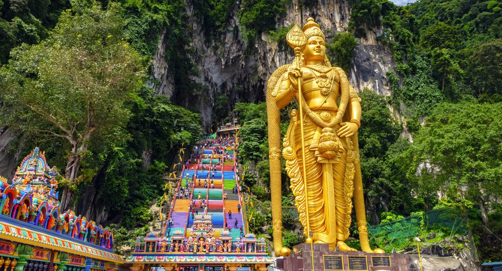 The iconic Batu Caves in Malaysia, featuring a vivid 272-step staircase leading into jungle-covered cliffs beneath a monumental golden deity.