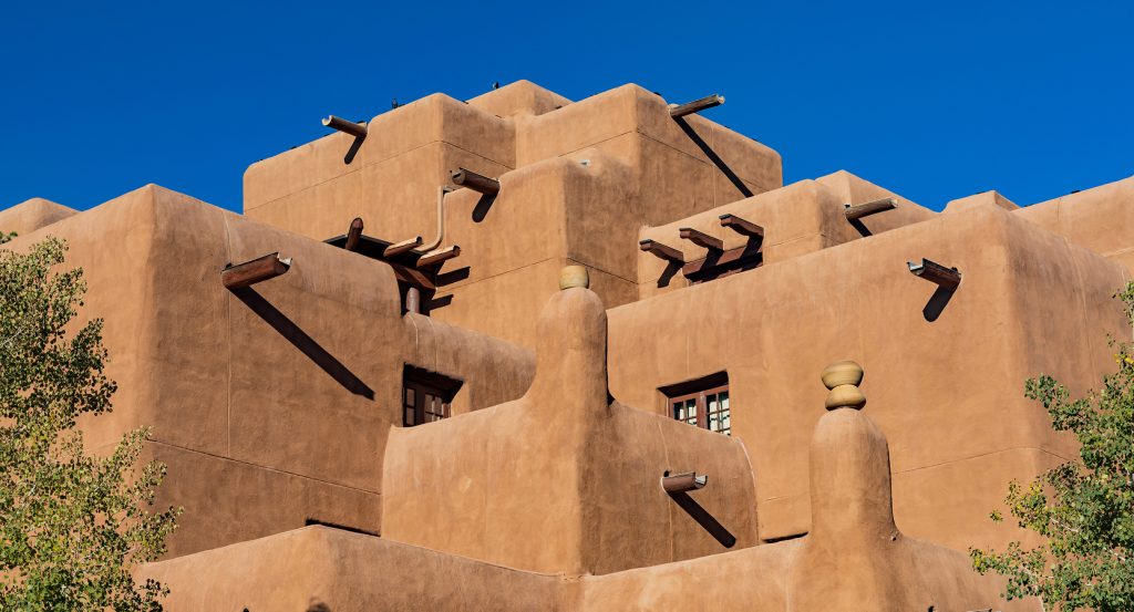 Tiered tan stucco walls and exposed wooden vigas of a classic Santa Fe adobe under clear blue skies.