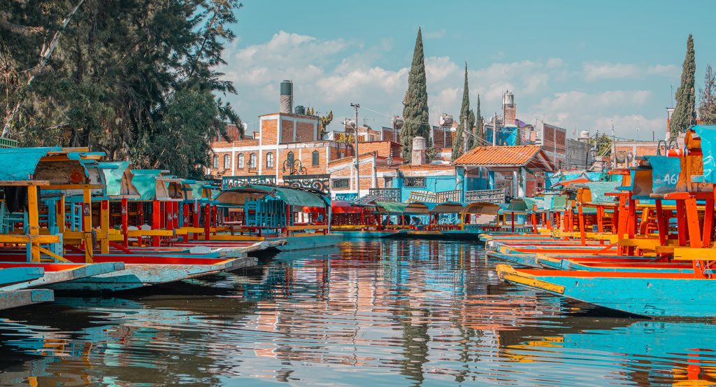 Traditional Mexican boats docked beside vibrant colonial homes and trees along Xochimilco’s waterways.