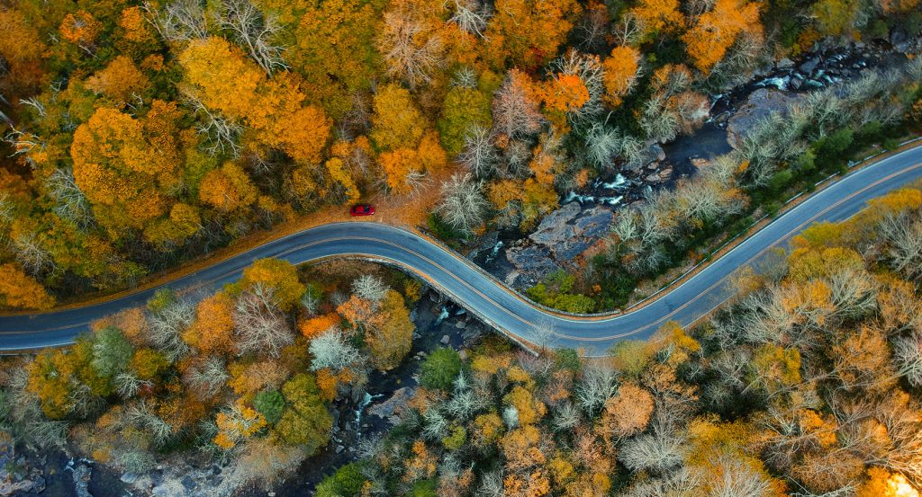 Aerial view of a winding mountain road in Asheville, North Carolina, surrounded by vibrant autumn foliage and a red car parked at a scenic overlook.