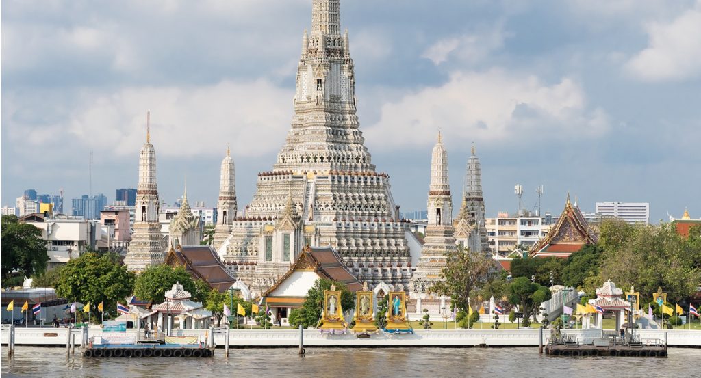 A riverside view of Wat Arun’s intricate architecture framed by fluttering flags and lush greenery in Bangkok, Thailand.