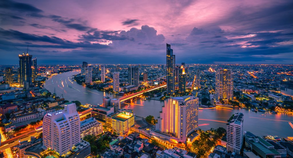 Twilight view of the Chao Phraya River winding through Bangkok, flanked by glittering high-rise hotels and glowing bridges under a purple sky.