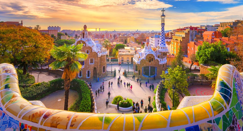 A sunrise view from Park Güell, Gaudí’s colorful tiled terrace curving into a skyline of terracotta rooftops and seaside light.