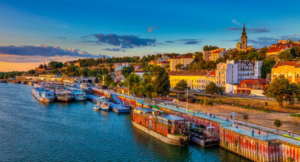 Evening view of Belgrade’s waterfront lined with boats, vibrant facades, and the Church of St. Michael in the distance.