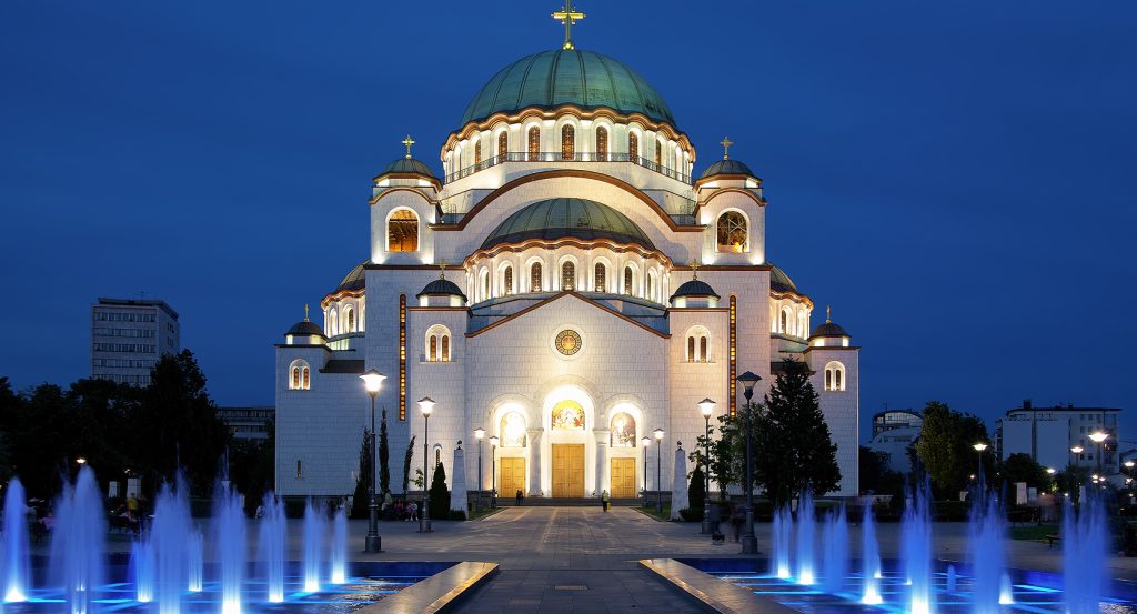 Nighttime view of the illuminated Church of Saint Sava in Belgrade, Serbia, with glowing blue fountains in the foreground and deep blue skies above.