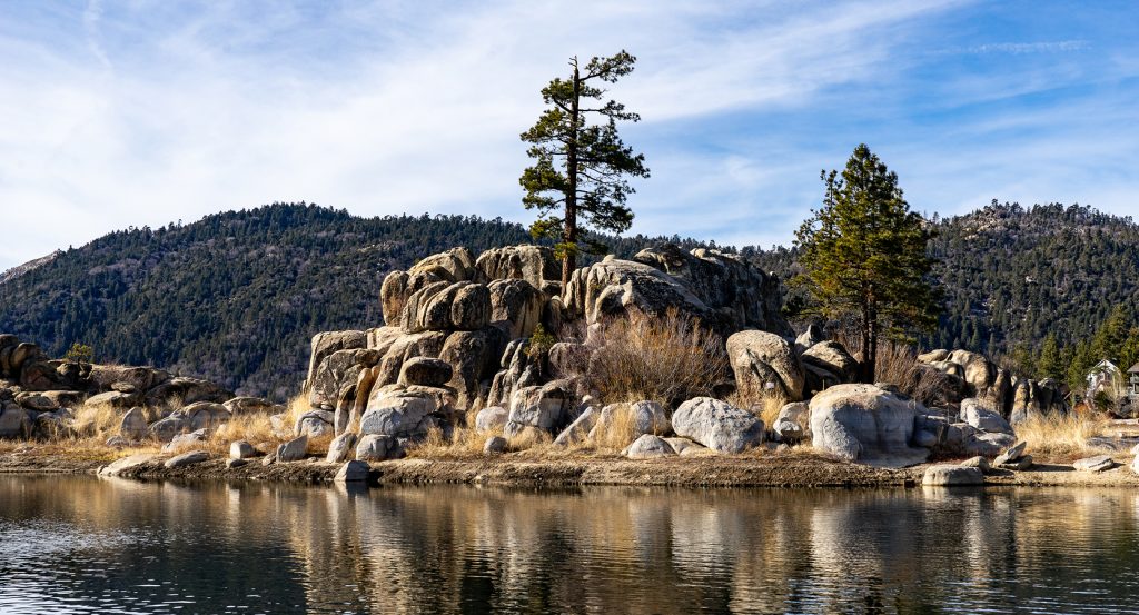 Tranquil scene at Big Bear Lake with boulders, evergreens, and clear blue skies mirrored in the water.