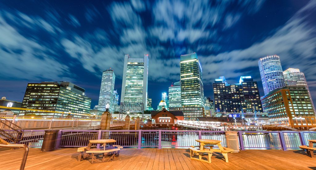 Boston harbor waterfront lit up at night, with iconic high-rises reflecting in the water and a cloudy sky overhead.