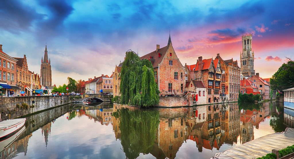 Charming canal houses and medieval towers in Brugge, Belgium, reflected in still waters at sunset with vibrant skies and gothic spires in the distance.