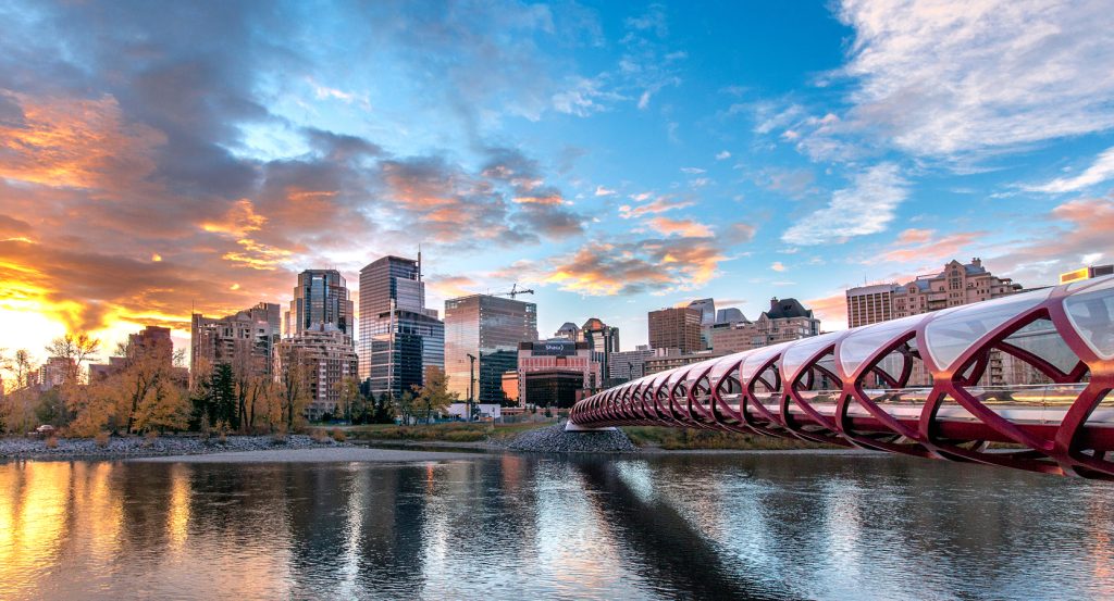 A glass-encased pedestrian bridge curves across the Bow River, leading into a modern skyline as the sun sets in vibrant oranges and blues.