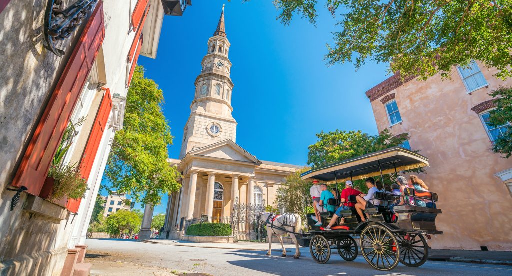 Tourists on a horse-drawn carriage ride passing by a historic church and pastel-colored buildings in Charleston, South Carolina.
