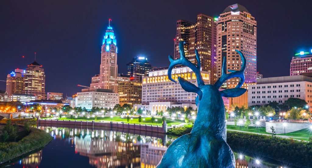 Nighttime view of downtown Columbus, Ohio, with a deer statue gazing at the glowing high-rises along the river.
