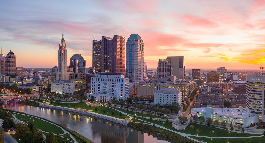 A scenic sunset casts a warm glow over the Columbus skyline, reflecting off the Scioto River with manicured green spaces lining the riverwalk.