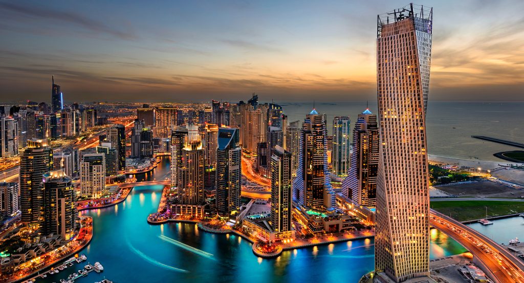A futuristic skyline of Dubai Marina glows at dusk, with the iconic twisting Cayan Tower rising above turquoise waters and luxury yachts below.