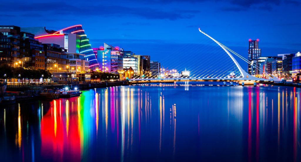 Reflection of city lights on the water in Dublin’s modern docklands district with iconic bridge and glowing skyline.
