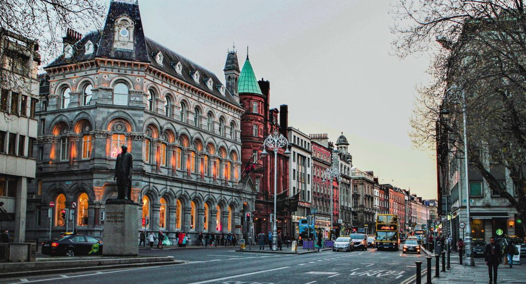 A bustling Dublin street lined with historic buildings and redbrick towers glows warmly at dusk as a double-decker bus rolls past.