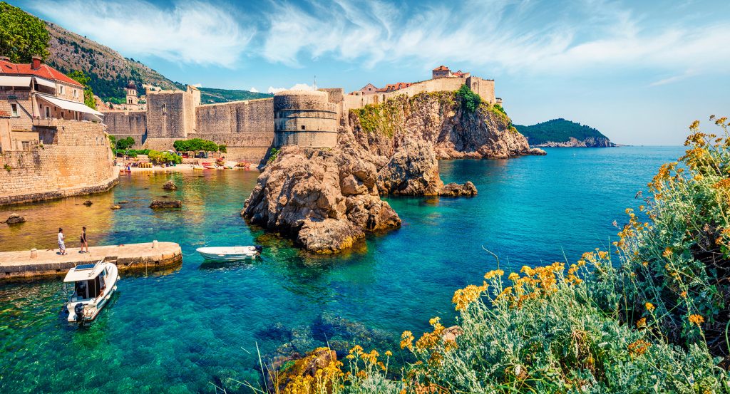 Scenic view of Dubrovnik’s Old Town walls rising above turquoise Adriatic waters with boats and rocky outcrops under a bright blue sky.