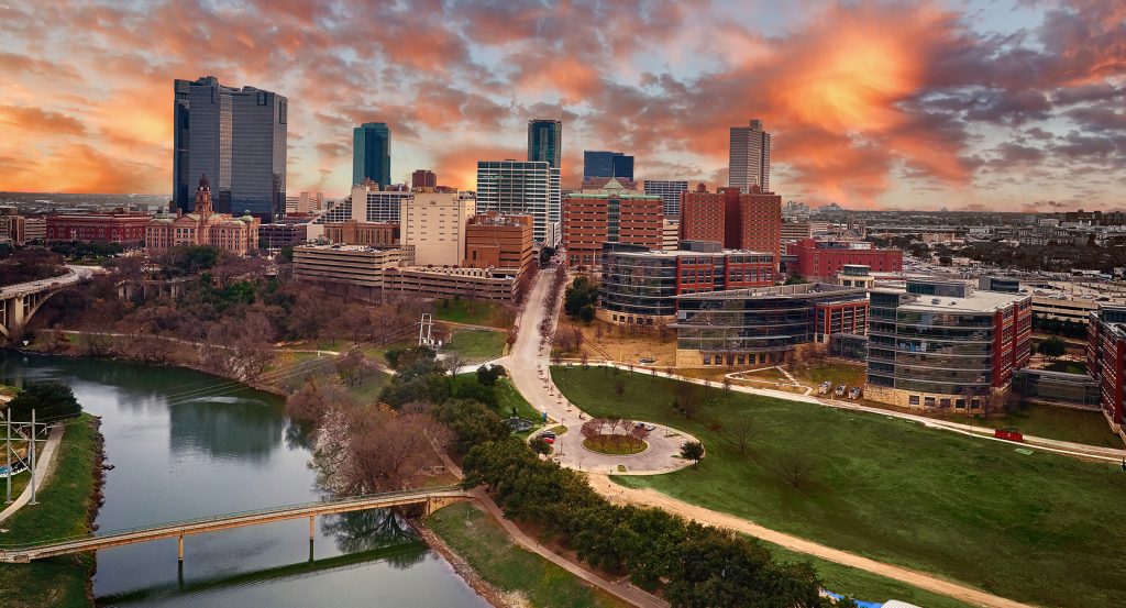 Downtown Fort Worth skyline at sunset with the Trinity River in the foreground and modern skyscrapers surrounding the historic Tarrant County Courthouse.
