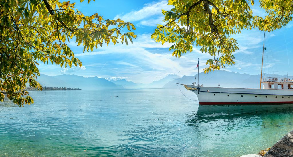 A peaceful white boat floats on the crystal-clear waters of Lake Geneva, framed by golden tree leaves and the distant silhouette of the Alps.