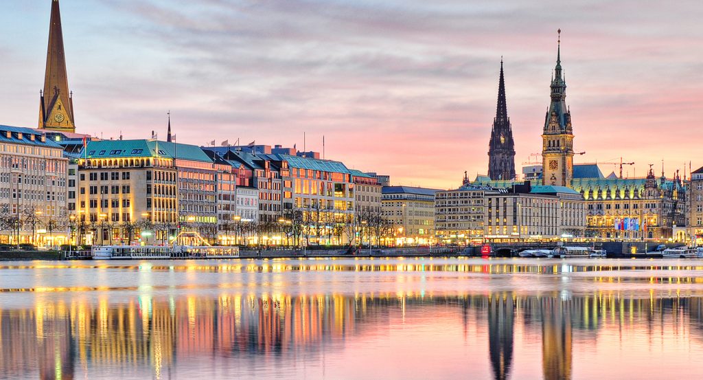 Illuminated buildings and church spires in Hamburg mirrored over tranquil evening waters.