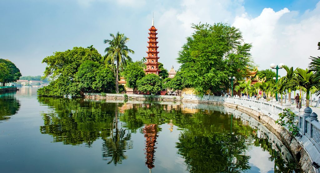 Reflections of Hanoi’s Tran Quoc Pagoda shimmer across a peaceful lakeside lined with trees and a stone path.