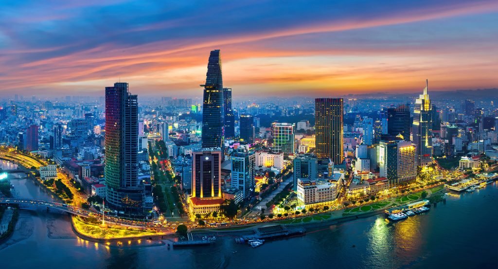 A vibrant aerial view of Ho Chi Minh City at dusk, showcasing the Saigon River, illuminated skyscrapers, and the Bitexco Financial Tower.