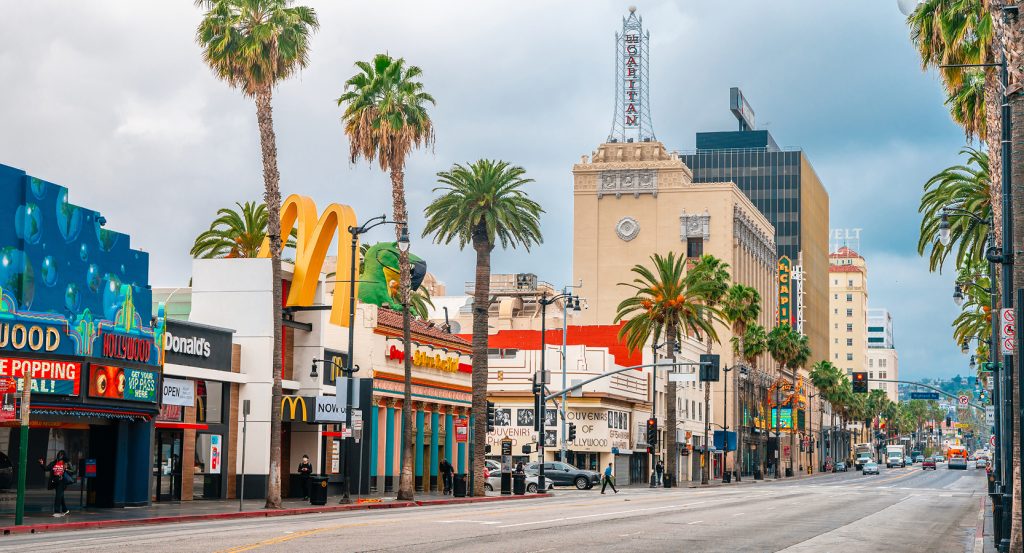 A lively street scene in Hollywood, Los Angeles, showcasing theaters, neon lights, and palm trees under a cloudy sky.