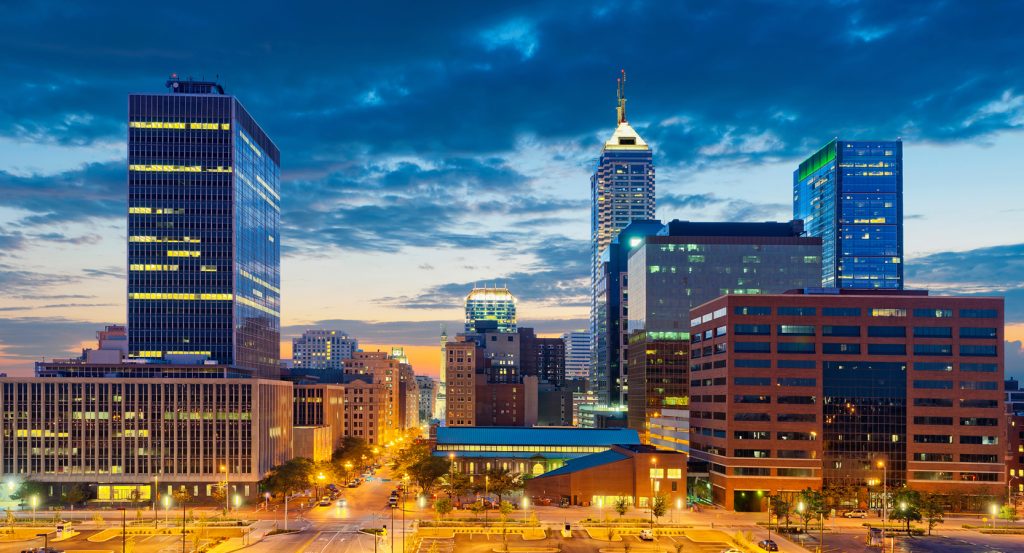 The skyline of downtown Indianapolis glows under a twilight sky, with tall modern buildings and warmly lit streets forming the city’s inviting core.