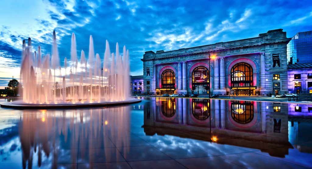 Kansas City’s Union Station and surrounding plaza lights up under a moody blue sunset with water reflections.