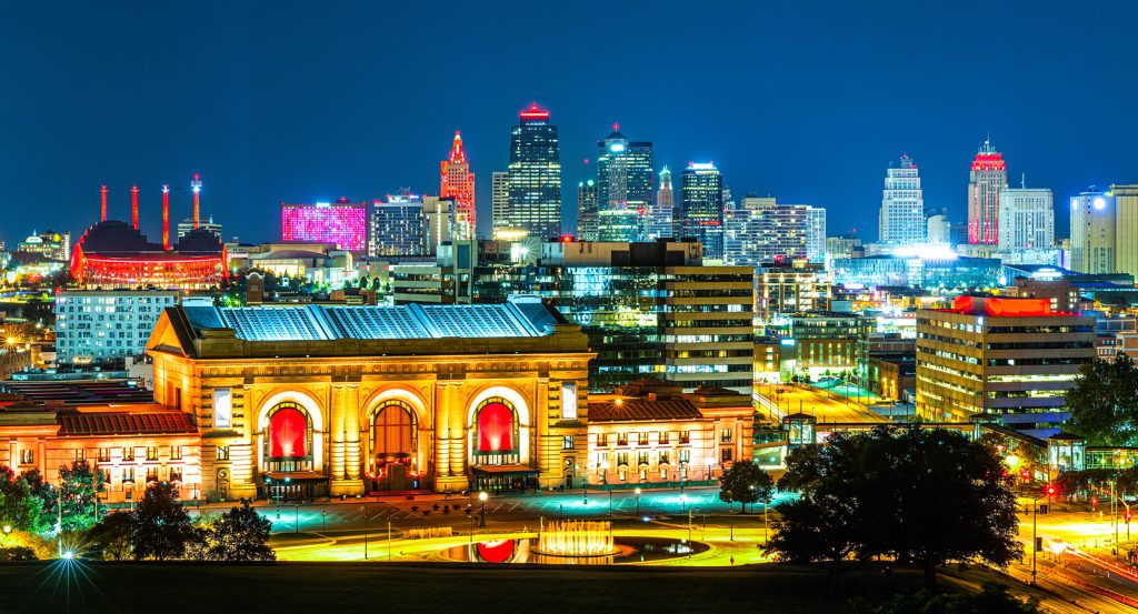 Nighttime view of Kansas City’s Union Station illuminated in golden hues, with the downtown skyline glowing in vibrant reds, purples, and neon lights.
