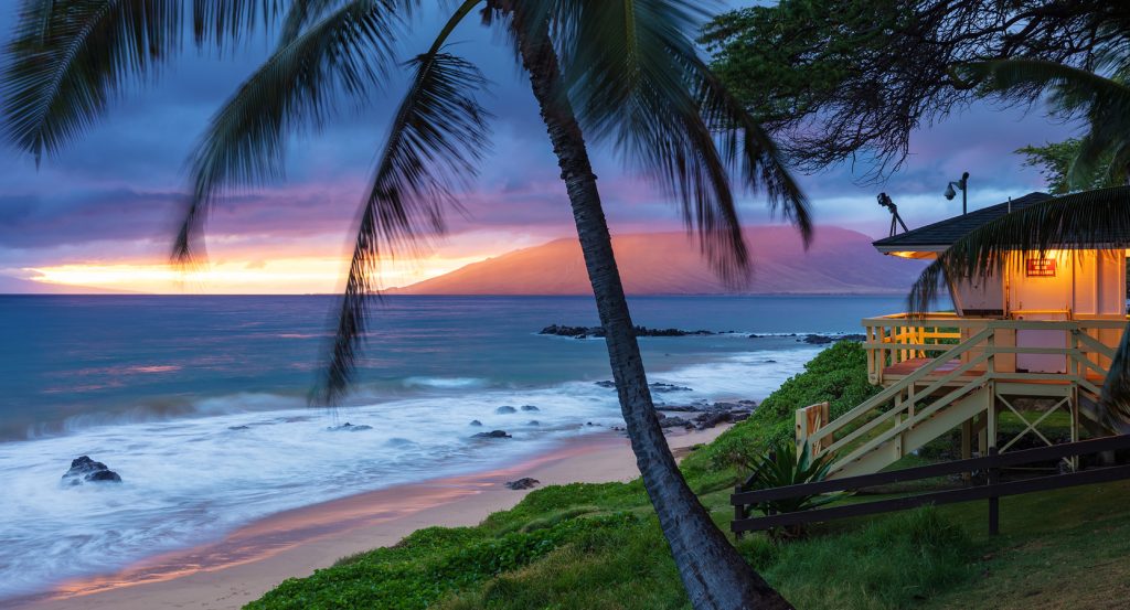 Golden hour over a tropical beach in Kihei, framed by lush palms and a cozy coastal house.