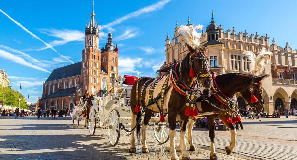 Decorated horses pull a white carriage through Krakow’s Rynek Główny under a bright blue sky