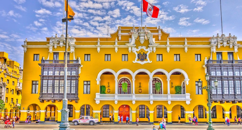 Vibrant colonial-style yellow building in Lima’s Plaza Mayor with ornate balconies, arches, and the Peruvian flag waving proudly above.