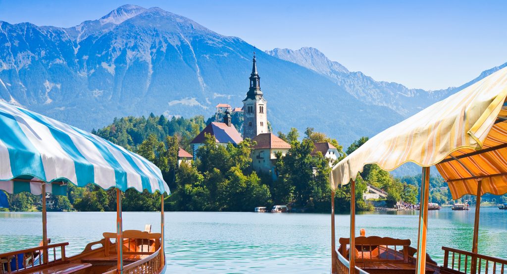 Colorful pletna boats frame a serene lake scene with a picturesque church and towering mountains in Slovenia.